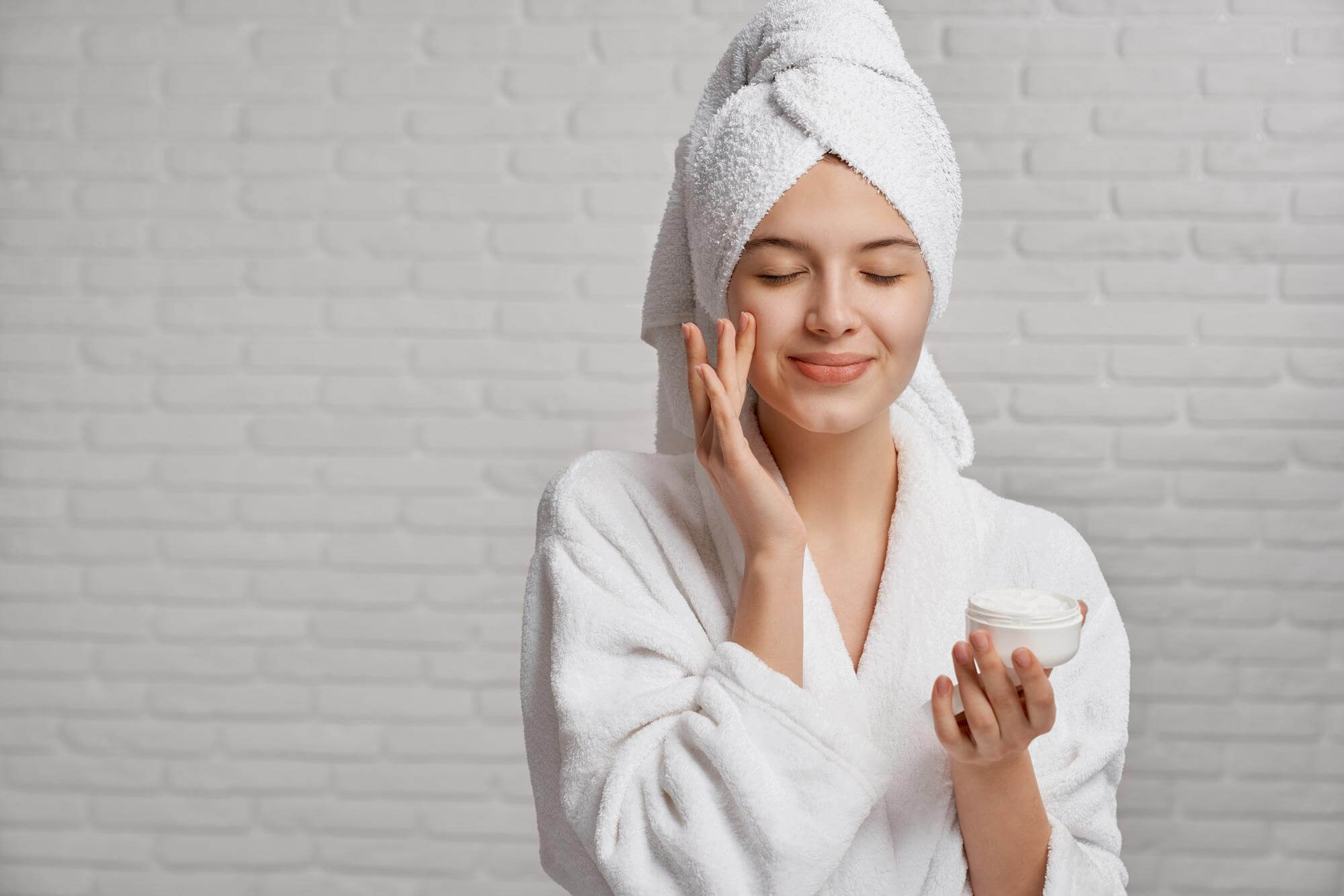 Woman in a white robe and towel wrapped around her head, holding a skincare product against a gray brick wall.